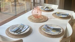 A dining table set for four with placemats and cutlery, a pink vase, and a view of an outdoor patio with chairs and a table. - The Deck @ VB - Linen, Wi-Fi, Pets, Secured Yard (Venus Bay)