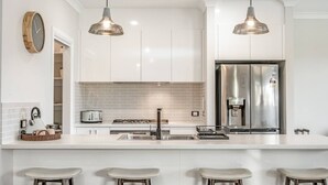 Een moderne keuken met witte kasten, roestvrijstalen apparatuur, hangende lampen en een groot eiland met barstoelen.