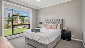 Een slaapkamer met een groot raam, een bed met een grijs hoofdeinde, een nachtkastje en een plafondventilator.