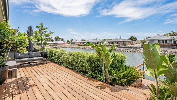 Een houten terras met een bank en plantenpotten, met uitzicht op een waterpartij en huizen.