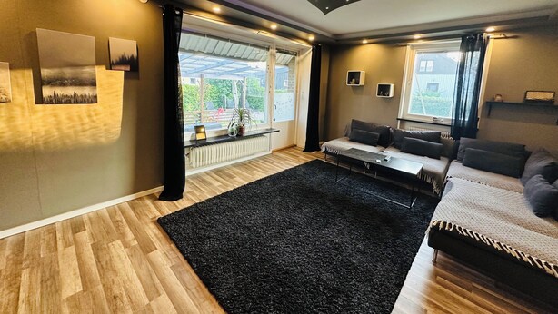 A modern living room with a sofa, a black rug, wooden flooring, and a view through a large window.