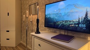 A room with a flat-screen TV displaying a mountain landscape, a white cabinet with drawers, a tall floor lamp, and a mirror on the wall.