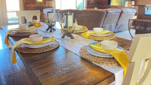 Une table de salle à manger en bois, mise avec de l’assiette blanche, des bols et des serviettes jaunes. On y trouve également des porte-bougies décoratifs et des sets de table en fibres tressées.