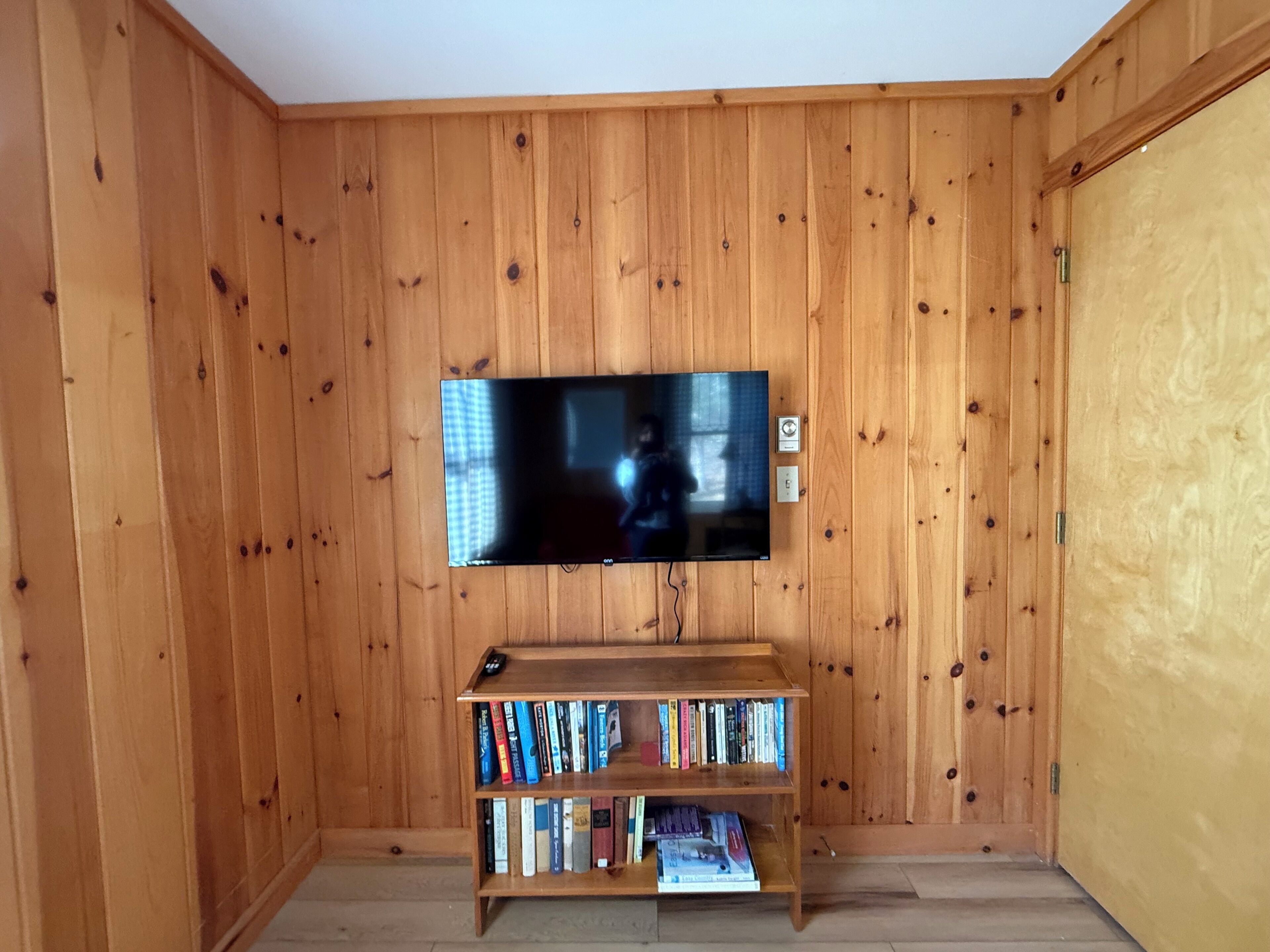 Una habitación con un revestimiento de madera en la pared, un televisor de pantalla plana montado sobre una estantería y una puerta a la derecha.