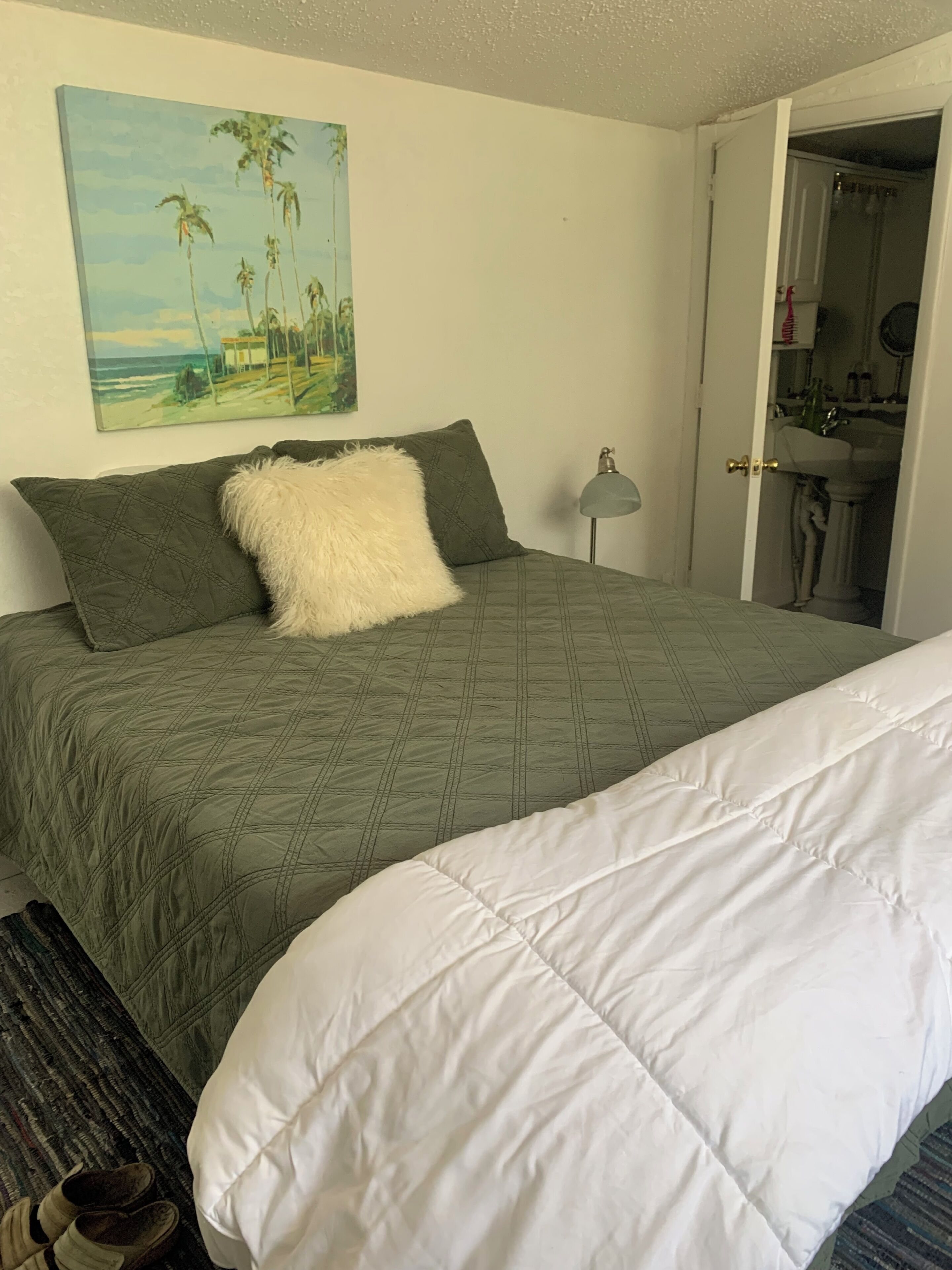A bed with a green quilt, a white pillow, and a painting of palm trees on the wall.