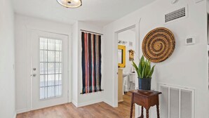 A hallway with a wooden floor, a door with a window, a striped curtain, a round woven wall hanging, and a potted plant on a small table.