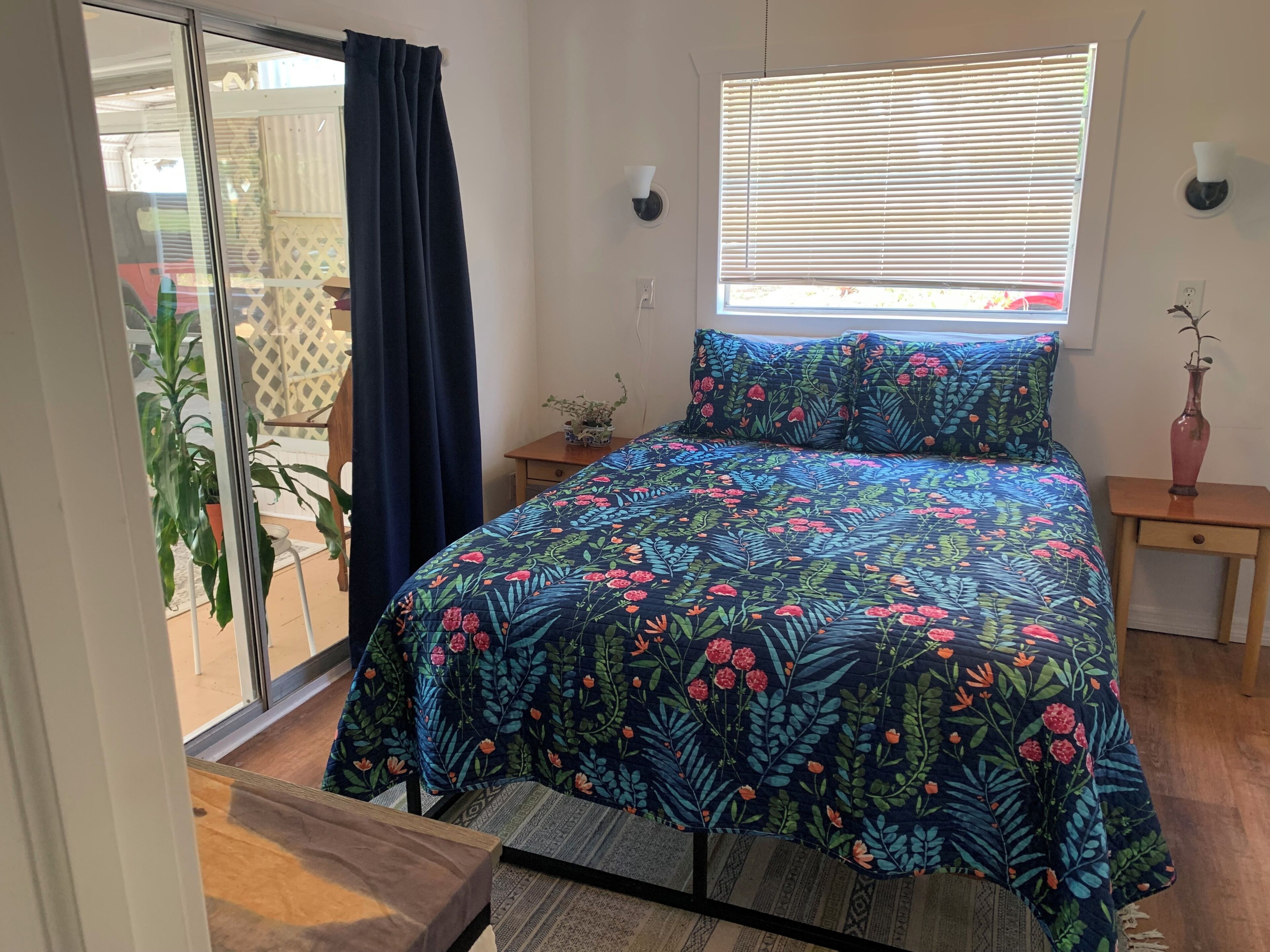 A bedroom with a bed, a window with blinds, a nightstand, a potted plant, and a sliding glass door leading to an outdoor area.