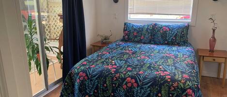 A bedroom with a bed, a window with blinds, a nightstand, a potted plant, and a sliding glass door leading to an outdoor area.