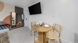 Una sala moderna con una mesa redonda de madera, sillas de mimbre, un televisor de pantalla plana montado en la pared y una puerta corrediza que da a otra habitación.