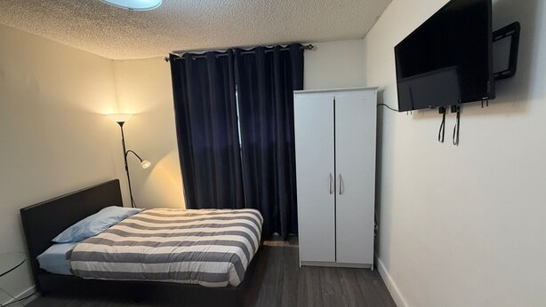 A bedroom with a bed, a wardrobe, a TV mounted on the wall, and a floor lamp. - Private Room Downtown with Kitchenette (Edmonton)