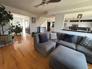 Ein modernes Wohnzimmer mit einem grauen Ecksofa, Holzboden und Deckenventilator.