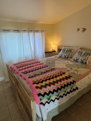 A bedroom with a wooden bed, a colorful blanket, a patterned quilt, a nightstand with a lamp, and a window with curtains.