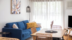 Ein Wohnzimmer mit blauem Sofa, einem Esstisch, Korbstühlen, einer Stehlampe und einem Bild an der Wand.