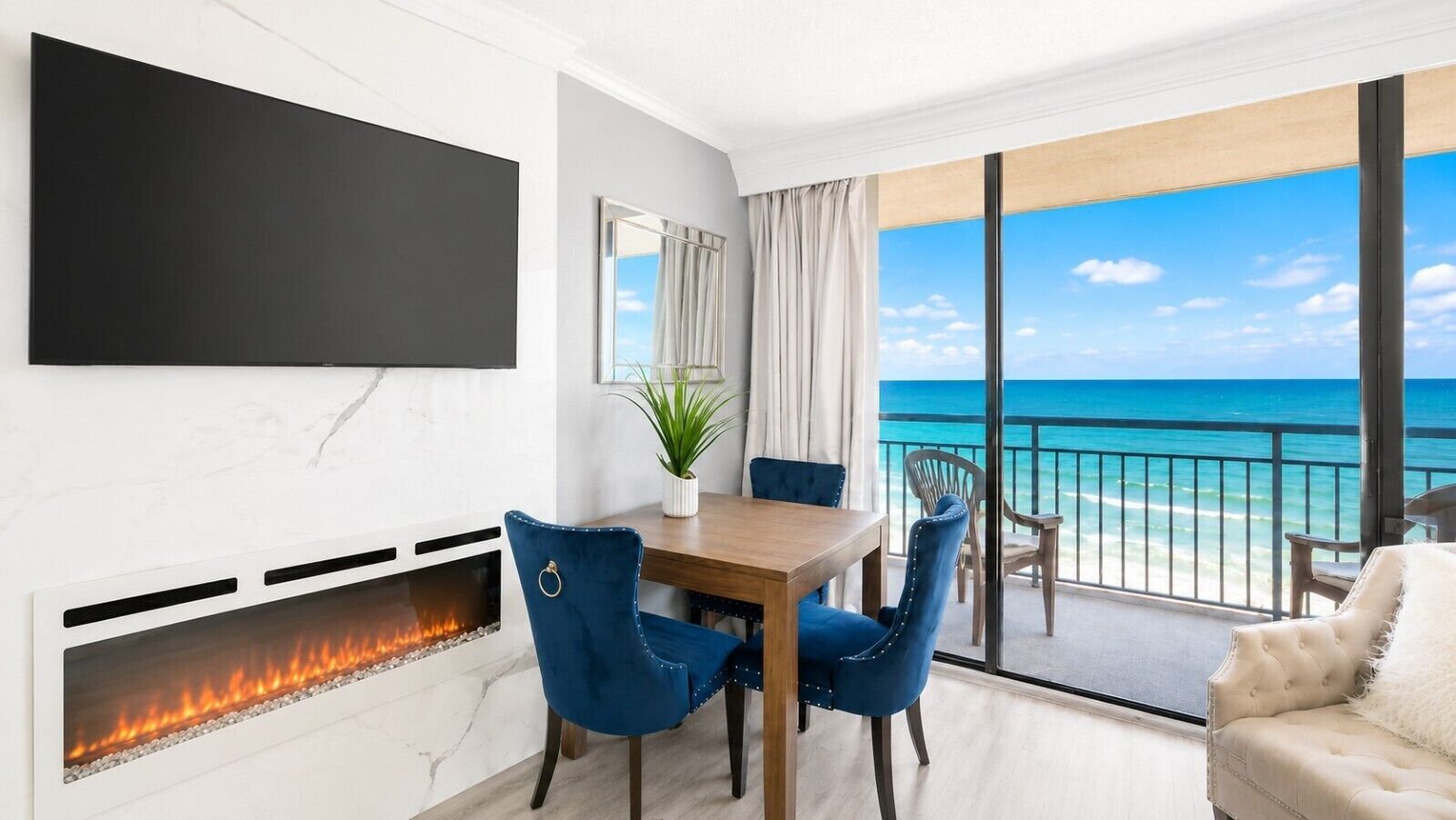 Una habitación moderna con chimenea, una mesa de comedor con sillas, un televisor en la pared y un balcón con vista al océano.