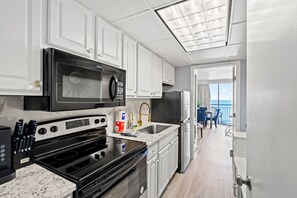 A modern kitchen with a microwave, stove, and sink, leading to a balcony with a view. - Oceanfront Condo with Resort Pools (Myrtle Beach)