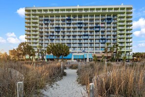 A multi-story hotel with a uniform exterior, a sandy pathway leading to the entrance, and a clear sky. - Oceanfront Condo with Resort Pools (Myrtle Beach)
