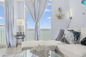 Ein modernes Wohnzimmer mit Meerblick, einem Glastisch, einer Couch mit Zierkissen, einem Beistelltisch mit Lampe und einer Wanddekoration in Form eines Ankers.