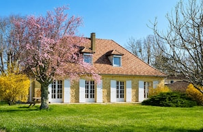 A stone house with a tiled roof, white shutters, and a large tree with pink blossoms in the front yard. - Beautiful riverside home on the Dordogne River (Eynesse)