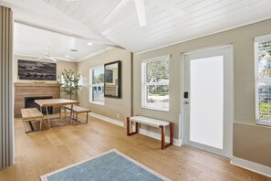 Una habitación amplia con suelo de madera, un banco, una mesa, una chimenea y ventanas grandes con persianas.