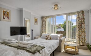 Ein Schlafzimmer mit Bett, Fernseher, Heizung, einem Fenster mit Vorhängen und Blick auf ein Haus.