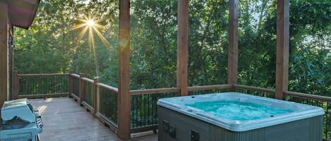 A hot tub on a wooden deck with a view of trees and the sun setting in the background.