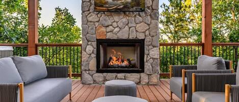 A stone fireplace with a fire, surrounded by outdoor furniture on a wooden deck.