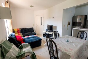 A living room with a sofa, ottoman, TV, dining table, chairs, and a kitchen area with a refrigerator. - Apto a 300 m da Praia do Sul com Piscina e Quadra (Ilhéus)