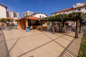 A courtyard with a tiled floor, outdoor seating, a pergola with vines, and surrounding buildings. - Apto a 300 m da Praia do Sul com Piscina e Quadra (Ilhéus)