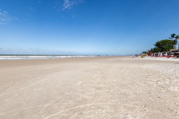 A wide sandy beach with a clear sky, a few people, and a row of buildings with thatched roofs on the right. - Apto a 300 m da Praia do Sul com Piscina e Quadra (Ilhéus)