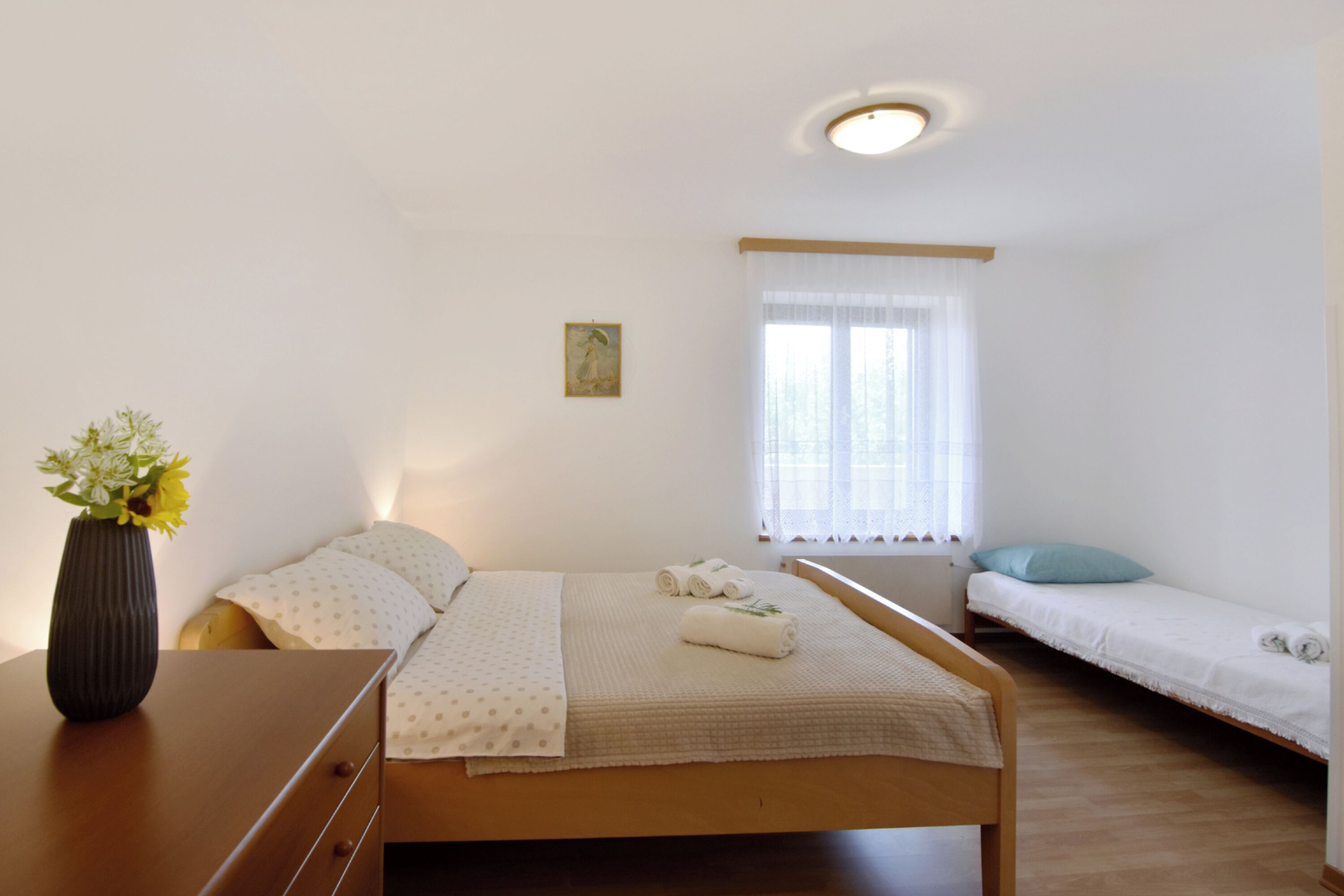 Une chambre à coucher avec un lit, un petit lit, une table de chevet, un vase avec des fleurs et une fenêtre avec des rideaux.