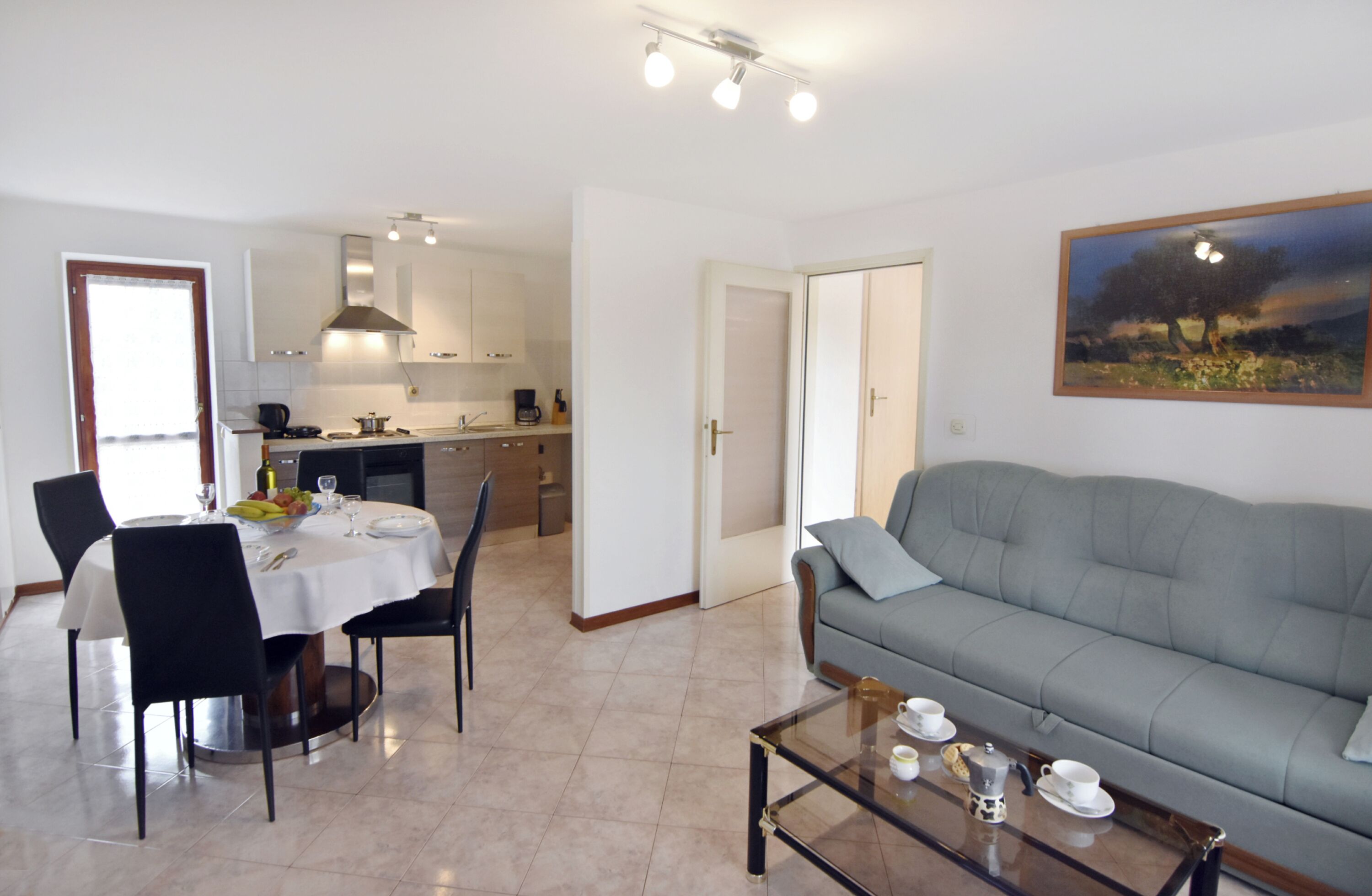 A living room with a sofa, a dining table with chairs, a kitchen in the background, and a painting on the wall.