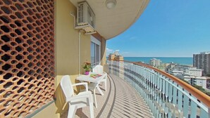 Varanda com mesa e cadeiras brancas, um vaso de planta e vista para o mar e edifícios.