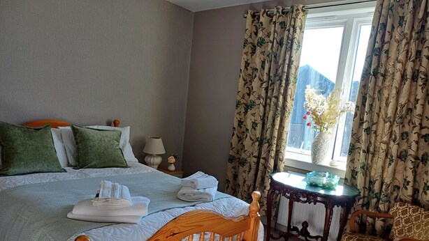 Una cama bien tendida con almohadas, una mesita de noche con lámpara, una silla y una ventana con cortinas.