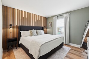 A modern bedroom with a large bed, wooden panel wall, bedside lamps, and a window with curtains. - Surf City Luxury Condo · Ocean Views · Pool (Surf City)