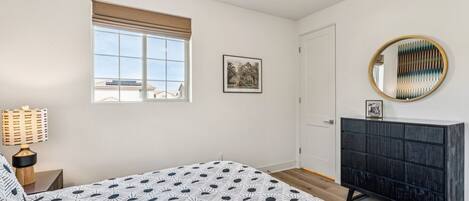 Een slaapkamer met een bed, een dressoir, een raam met jaloezieën, een fotokader aan de muur en een plafondlamp.