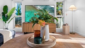 A modern living room with a wooden coffee table, a vase with flowers, a candle, and a flat-screen TV displaying a tropical beach scene. - Cullen Bay Retreat | Pool, Balcony & Marina Stroll (Larrakeyah)
