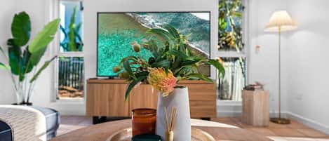 A modern living room with a wooden coffee table, a vase with flowers, a candle, and a flat-screen TV displaying a tropical beach scene.