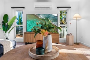 Ein modernes Wohnzimmer mit Couchtisch aus Holz, einer Blumenvase, einer Kerze und einem Flachbildfernseher, der eine tropische Strandlandschaft zeigt.