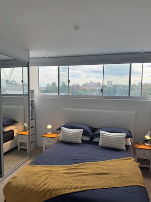 A bedroom with a bed, bedside tables, a mirror, and a view of the city. - Golden Mile Beachside (Manly)