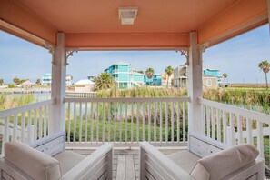 A covered outdoor seating area with white railings and beige cushions, overlooking a grassy area with buildings in the background. - Four Winds | Pond Views + Coastal Charm! (Port Aransas)