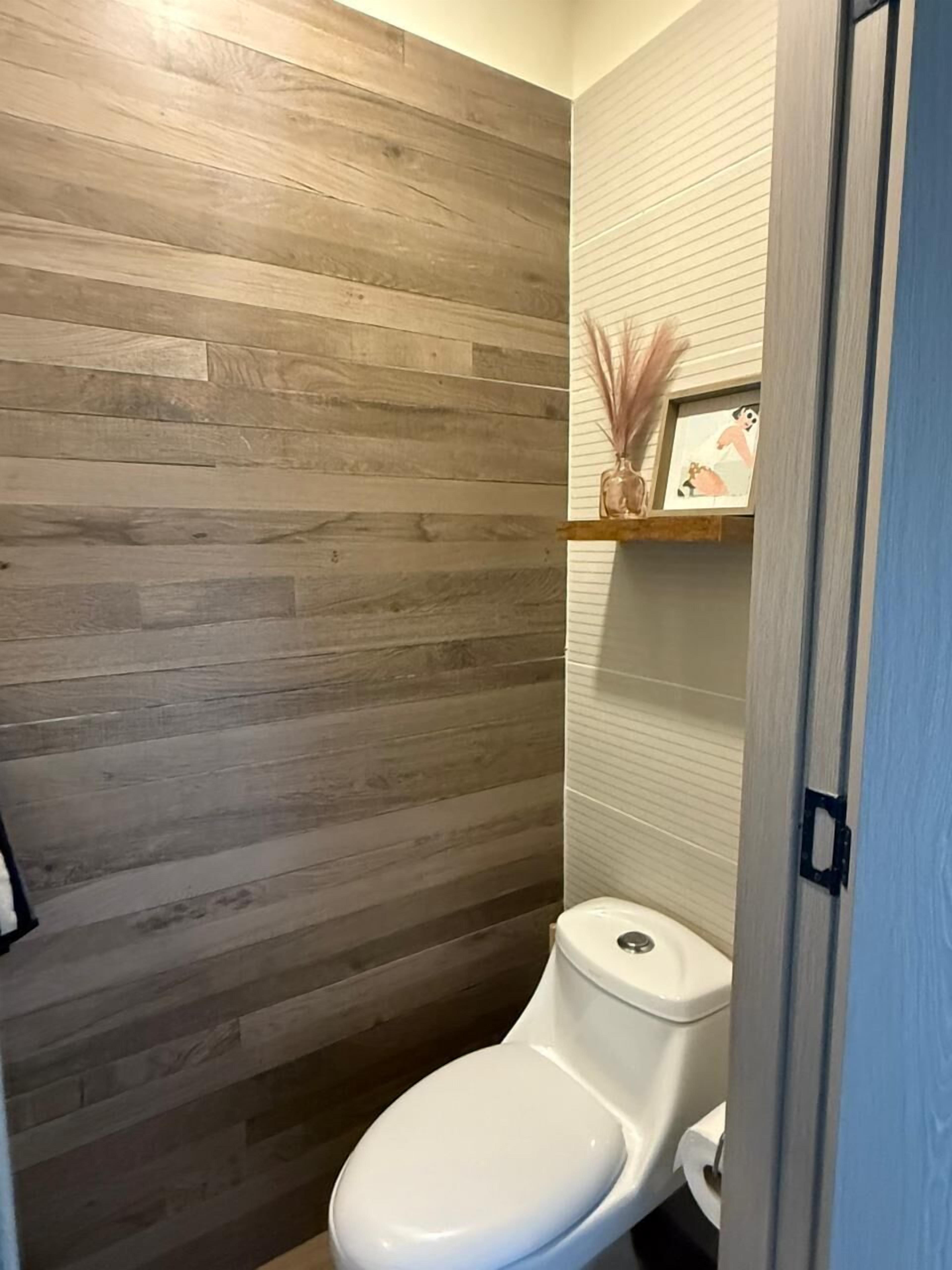 A bathroom with a wooden wall, a white toilet, and a shelf with a vase and picture.