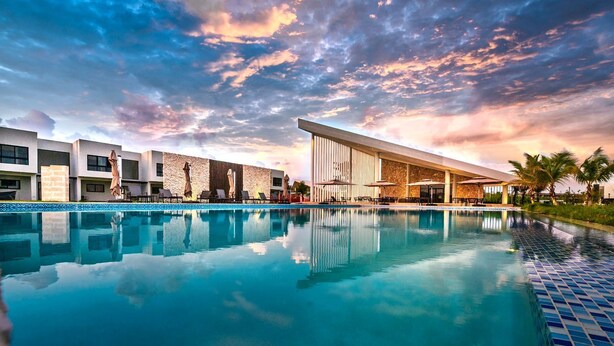 A swimming pool with lounge chairs and umbrellas, modern buildings with large windows, and a sunset sky. - Cozy 1-bedroom condo with AC in welcoming Punta Cana (Punta Cana)