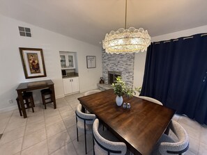 Un comedor con una mesa de madera, sillas, una araña, una chimenea y una cocina al fondo.