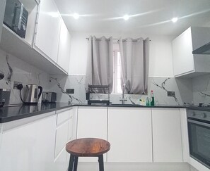Eine Küche mit weißen Schränken, einem Holzhocker, einem Wasserkocher, einem Toaster und einem Fenster mit Vorhängen.