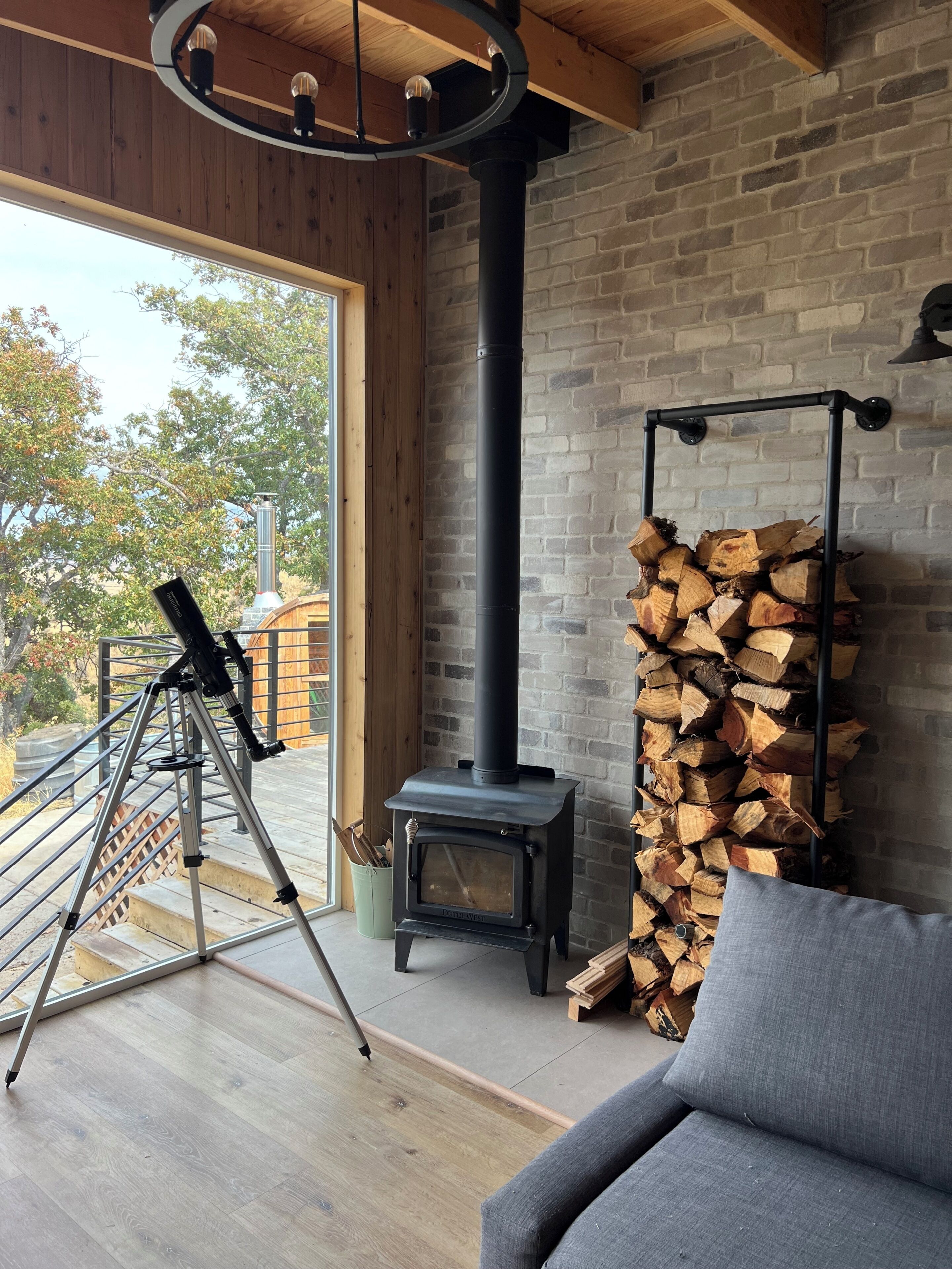 A modern interior with a wood-burning stove, a telescope on a tripod, and a stack of firewood against a brick wall.