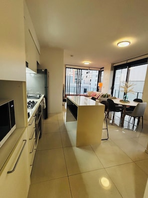 A modern kitchen with a central island, a dining area with a table and chairs, and a view of buildings through large windows. - Listen to & watch the waves, swim, surf, see dolphins & whales! (Newcastle)