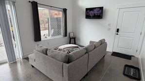 Un salon moderne avec un canapé d’angle gris, un téléviseur à écran plat fixé au mur et une grande fenêtre avec des rideaux.