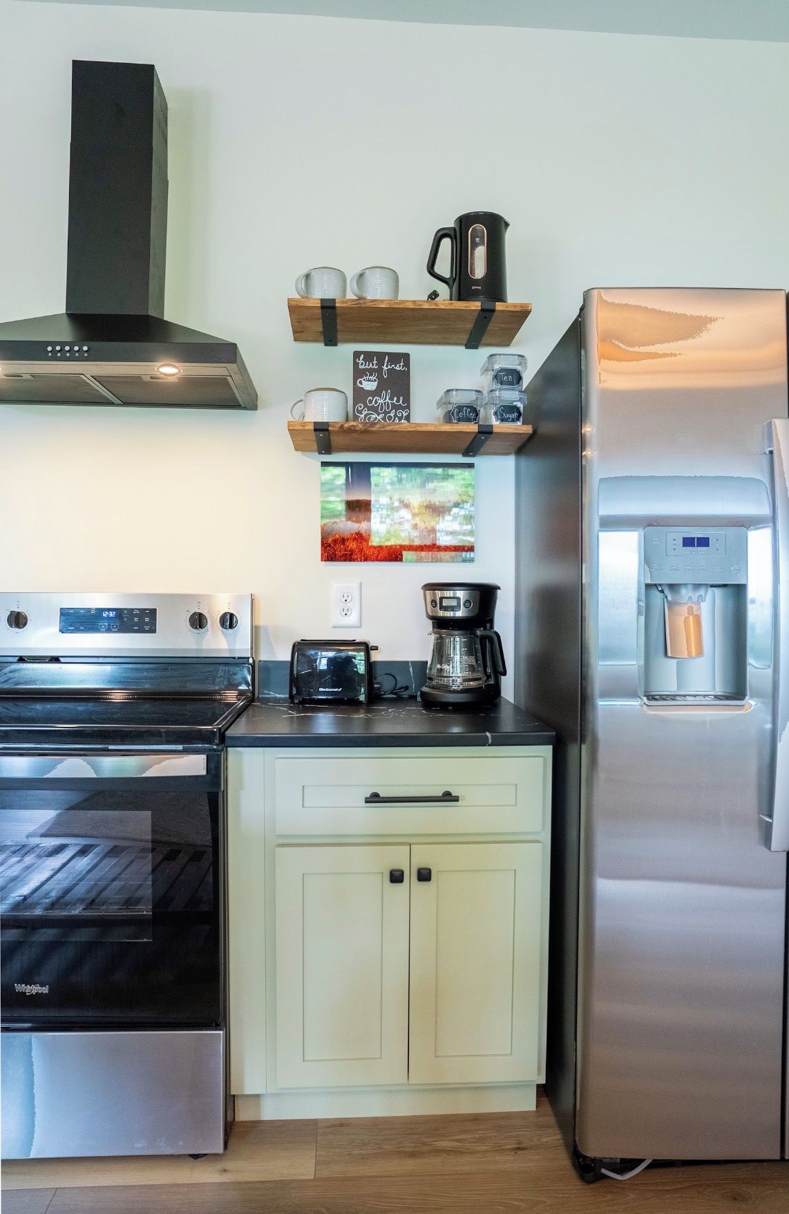Eine kleine Küche mit einem Edelstahl-Kühlschrank, einem Einbauofen, einer Kaffeemaschine und einem Regal mit Gläsern und einem Schild.