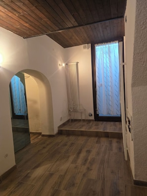 A room with a wooden floor, white walls, a radiator, a chair, and a window with curtains. - Duplex Apartment (Bologna)