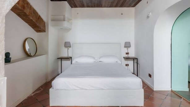 A bedroom with a wooden ceiling, white walls, a bed with white bedding, two bedside tables with lamps, and a circular mirror on the wall. - Duplex Apartment (Bologna)